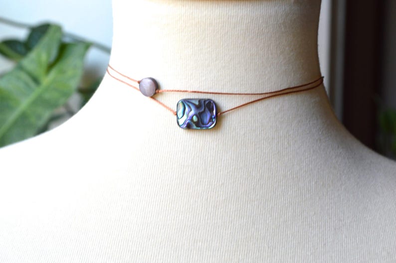 May include: Abalone shell choker necklace. The necklace features a rectangular abalone shell pendant with iridescent colors of blue, green, and purple. A small, round, gray bead is on the side. The necklace is displayed on a mannequin.