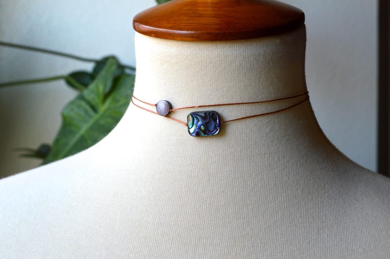 May include: A delicate choker necklace with two strands. One strand features a small, round, gray bead, and the other has a rectangular abalone shell pendant. The necklace is displayed on a mannequin.