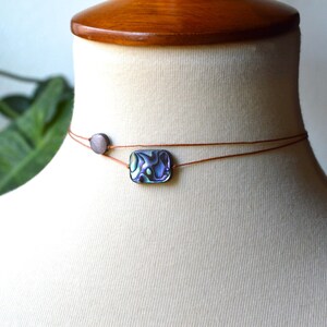May include: Abalone shell choker necklace with a rectangular pendant and a small round bead. The necklace is strung on a thin, brown cord and displayed on a mannequin. The abalone shell pendant has iridescent colors.