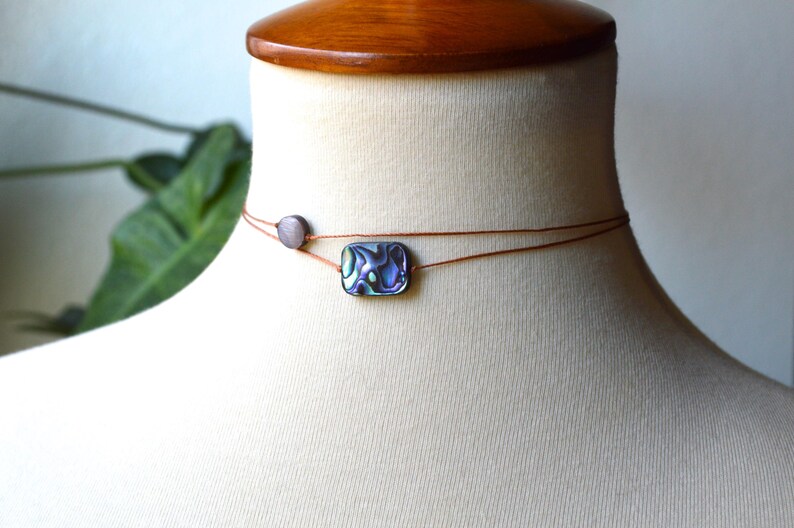 May include: Abalone shell choker necklace with a rectangular pendant and a small round bead. The necklace is strung on a thin, brown cord and displayed on a mannequin. The abalone shell pendant has iridescent colors.