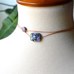 May include: Abalone shell choker necklace. The necklace features a small, rectangular abalone shell pendant with iridescent colors of blue, green, and purple. The pendant is strung on a thin, brown cord, with a small, round bead.