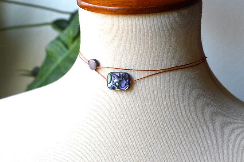 May include: Abalone shell choker necklace. The necklace features a small, rectangular abalone shell pendant with iridescent colors of blue, green, and purple. The pendant is strung on a thin, brown cord, with a small, round bead.