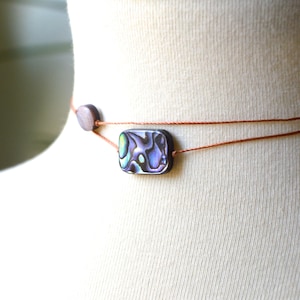 May include: Abalone shell necklace with a rectangular pendant and a small round bead. The necklace features a double strand of brown cord. The pendant has iridescent blue, green, and purple hues.