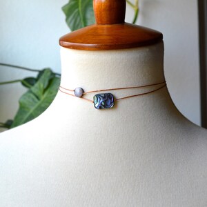 May include: A delicate choker necklace featuring two strands of thin, brown cord. The top strand holds a small, round, light-colored bead, while the bottom strand showcases a rectangular, iridescent abalone shell pendant. The necklace is displayed on a mannequin.