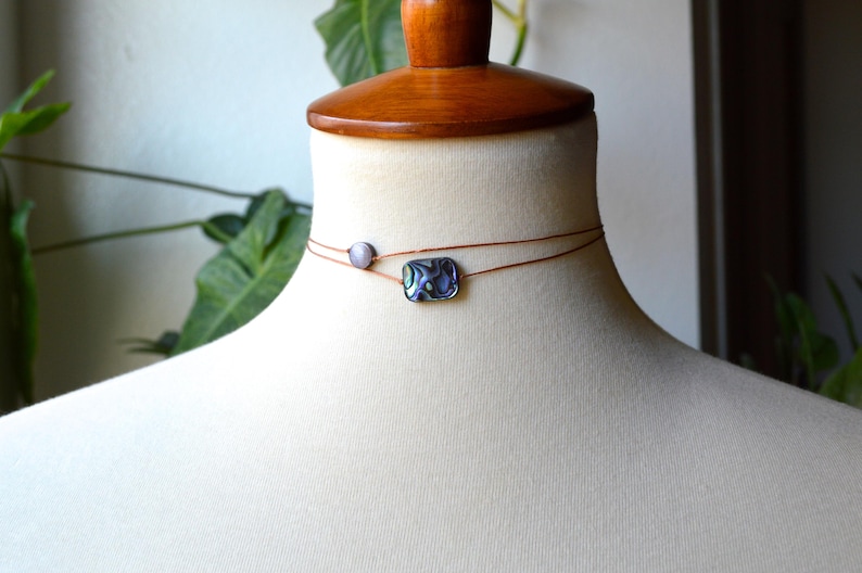 May include: A delicate choker necklace featuring two strands of thin, brown cord. The top strand holds a small, round, light-colored bead, while the bottom strand showcases a rectangular, iridescent abalone shell pendant. The necklace is displayed on a mannequin.