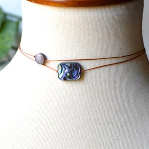 May include: A delicate choker necklace featuring a rectangular abalone shell pendant with iridescent colors of blue, green, and purple. The necklace is strung on two thin, brown cords, with a small, round, gray bead accent. The necklace is displayed on a mannequin.