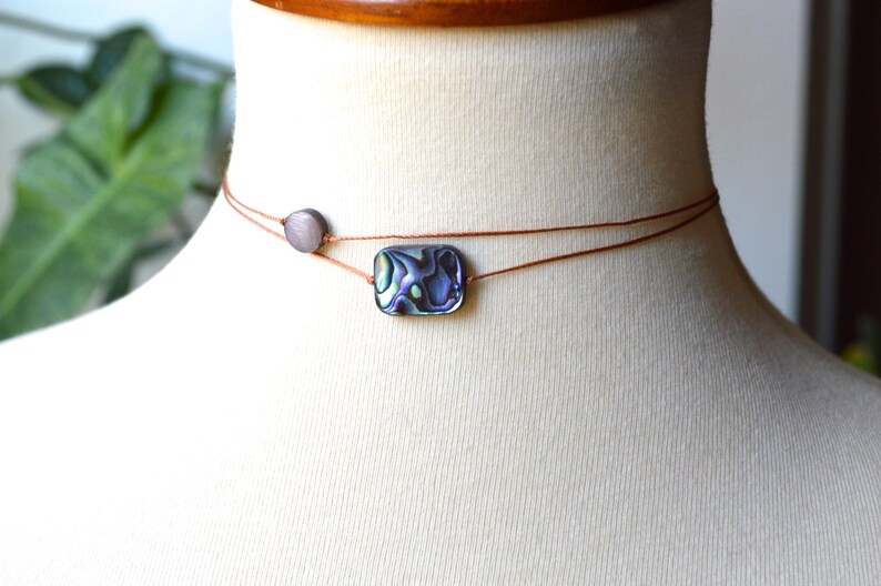 May include: A delicate choker necklace featuring a rectangular abalone shell pendant with iridescent colors of blue, green, and purple. The necklace is strung on two thin, brown cords, with a small, round, gray bead accent. The necklace is displayed on a mannequin.