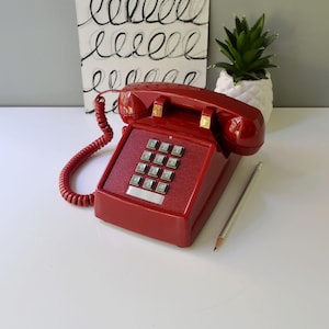 May include: A vintage red telephone with a push-button keypad. The phone has a coiled handset cord and a rectangular base. A silver pencil rests beside the phone. In the background, there is a black and white abstract art piece and a small potted plant.