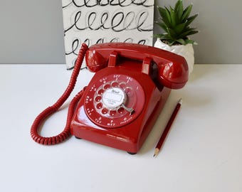 Puede incluir: Un teléfono rojo vintage con dial giratorio y cable de auricular en espiral. El teléfono tiene un dial circular con números y letras. Un lápiz descansa junto al teléfono. Una pequeña planta suculenta en una maceta blanca y una obra de arte abstracta en el fondo.