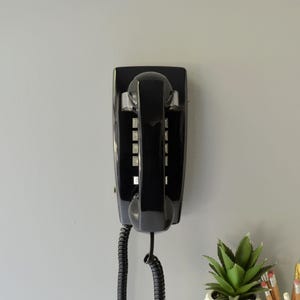 May include: A glossy black wall-mounted telephone featuring a rectangular base and a matching handset resting in the center. The device includes a vertical numeric keypad with silver-colored buttons and a coiled black cord extending from the bottom.