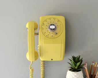 Puede incluir: Un teléfono de pared giratorio de color amarillo brillante que cuenta con un dial circular con números y letras, un auricular a juego en el lateral y un cable en espiral que cuelga hacia abajo. El dispositivo tiene un acabado de plástico liso.