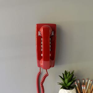 May include: A bright red, vintage wall-mounted telephone. The phone has a handset, a dial pad, and a coiled cord. The phone is mounted on a gray wall. A small succulent plant in a white pot and pencils are in the foreground.