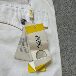 May include: Close-up of off-white Versace jeans with a pocket and a silver button. A yellow and white tag with "VERSACE JEANS COUTURE" is attached. The tag has a silver medallion and text.