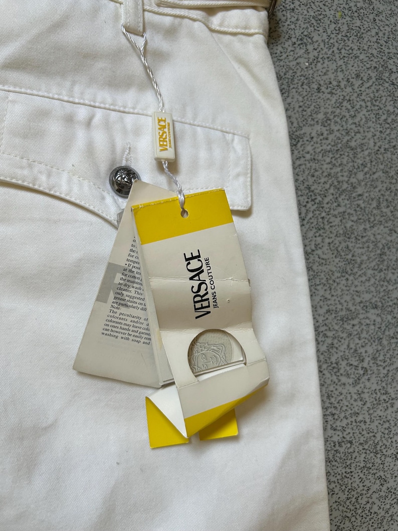 May include: Close-up of off-white Versace jeans with a pocket and a silver button. A yellow and white tag with "VERSACE JEANS COUTURE" is attached. The tag has a silver medallion and text.