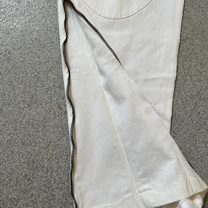 May include: Close-up of white pants with a black zipper detail running along the side. The pants have a textured, stitched pattern and a circular patch on the back. The zipper is partially open, revealing the interior fabric.