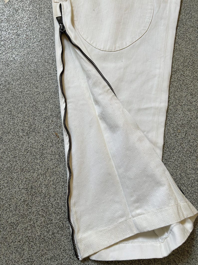 May include: Close-up of white pants with a black zipper detail running along the side. The pants have a textured, stitched pattern and a circular patch on the back. The zipper is partially open, revealing the interior fabric.