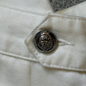 May include: Close-up of a white garment with a silver button. The button is round with a detailed embossed design and the words "JEANS COUTURE VERSACE" around the edge. The fabric has visible stitching.