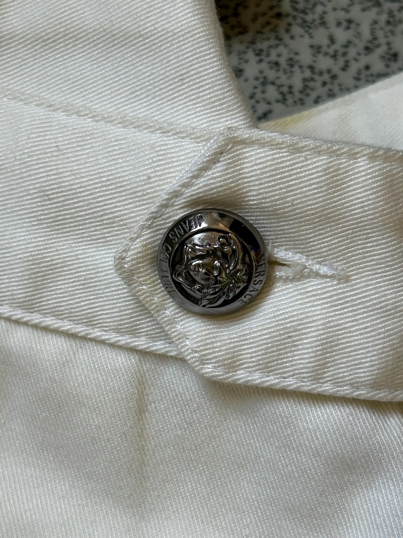 May include: Close-up of a white garment with a silver button. The button is round with a detailed embossed design and the words "JEANS COUTURE VERSACE" around the edge. The fabric has visible stitching.