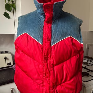 May include: A red and blue corduroy vest with a high collar. The vest has a quilted design with a red body and a blue corduroy collar and shoulders. White piping accents the design. The vest buttons up the front.