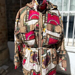Peut inclure: Une veste à manches longues arborant un imprimé géométrique et abstrait multicolore dans des tons magenta, beige, blanc et vert foncé. La veste comprend une fermeture éclair frontale, une taille à cordon, une capuche et deux poches poitrine.