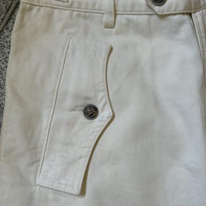 May include: Close-up of off-white pants with a buttoned pocket and decorative silver buttons. The pants have a high waist and a tailored fit, showcasing a classic design. The fabric appears to be a sturdy cotton or blend, suitable for various occasions.