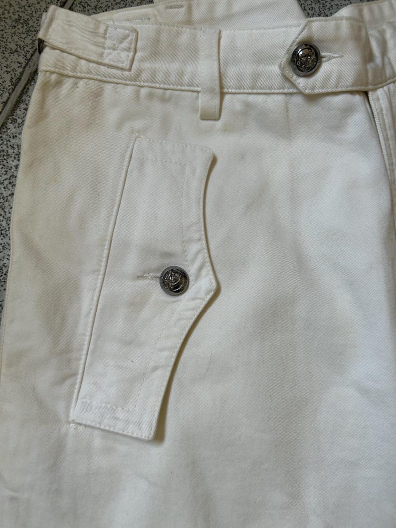 May include: Close-up of off-white pants with a buttoned pocket and decorative silver buttons. The pants have a high waist and a tailored fit, showcasing a classic design. The fabric appears to be a sturdy cotton or blend, suitable for various occasions.
