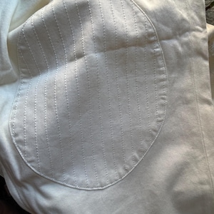 May include: Close-up of a cream-colored fabric with a circular patch. The patch features vertical stitched lines, creating a textured pattern. The fabric appears to be a light, possibly cotton or linen material.