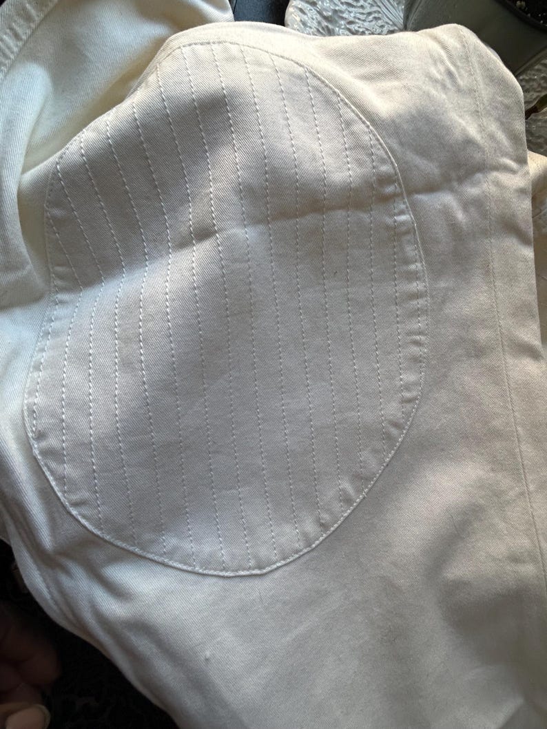 May include: Close-up of a cream-colored fabric with a circular patch. The patch features vertical stitched lines, creating a textured pattern. The fabric appears to be a light, possibly cotton or linen material.
