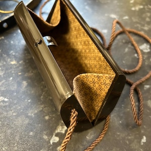 May include: A dark brown, hard-shell clutch bag with a smooth, glossy exterior and a hinged opening. The interior is lined with a tan fabric featuring a repeating brown monogram pattern. A thin, twisted brown rope cord serves as a shoulder strap.