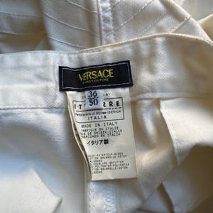 May include: Close-up of a pair of off-white Versace jeans. The label is visible, showing the brand name and size information. The fabric has a textured, ribbed design.