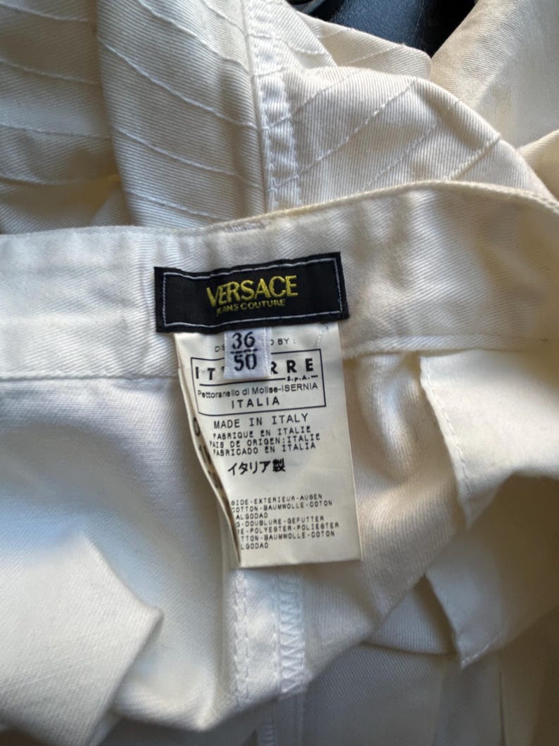 May include: Close-up of a pair of off-white Versace jeans. The label is visible, showing the brand name and size information. The fabric has a textured, ribbed design.