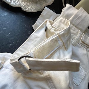 May include: Close-up of off-white pants with a matching belt. The belt has a black metal ring and a velcro closure. The pants have a pocket and a structured waistband. The fabric appears to be a sturdy cotton or linen blend.