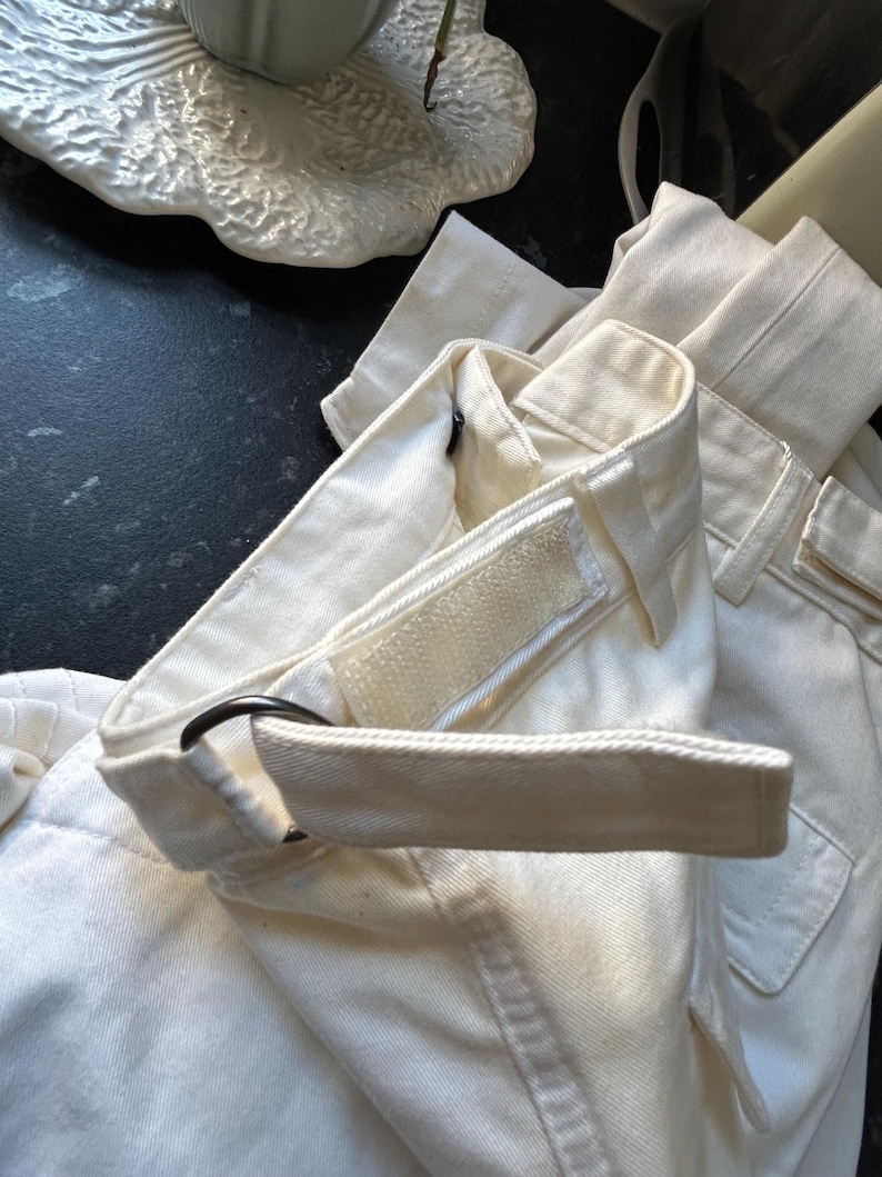 May include: Close-up of off-white pants with a matching belt. The belt has a black metal ring and a velcro closure. The pants have a pocket and a structured waistband. The fabric appears to be a sturdy cotton or linen blend.
