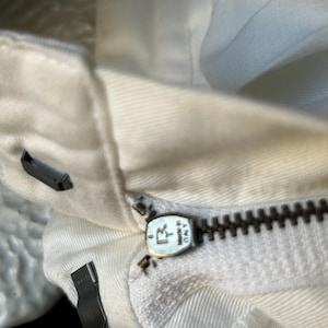 May include: Close-up of a white garment with a visible zipper. The zipper is dark brown and has a metal pull tab with the text "MADE IN ITALY". The fabric is a light, off-white color.