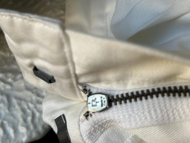 May include: Close-up of a white garment with a visible zipper. The zipper is dark brown and has a metal pull tab with the text "MADE IN ITALY". The fabric is a light, off-white color.