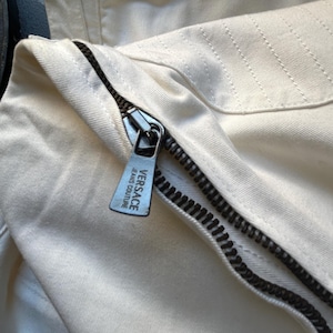 May include: Close-up of cream-colored denim fabric with a black zipper. The zipper pull features the "VERSACE JEANS COUTURE" logo. The fabric has visible stitching and a metal ring detail.