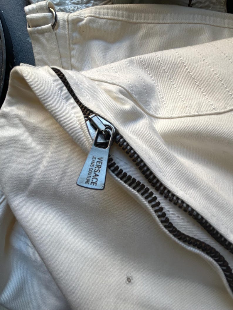 May include: Close-up of cream-colored denim fabric with a black zipper. The zipper pull features the "VERSACE JEANS COUTURE" logo. The fabric has visible stitching and a metal ring detail.