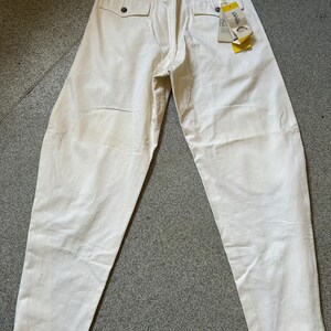 May include: Off-white pants with a relaxed fit, featuring two back pockets with button closures and a belt. A tag with the brand name is attached. The pants are made of a sturdy fabric, suitable for casual wear.