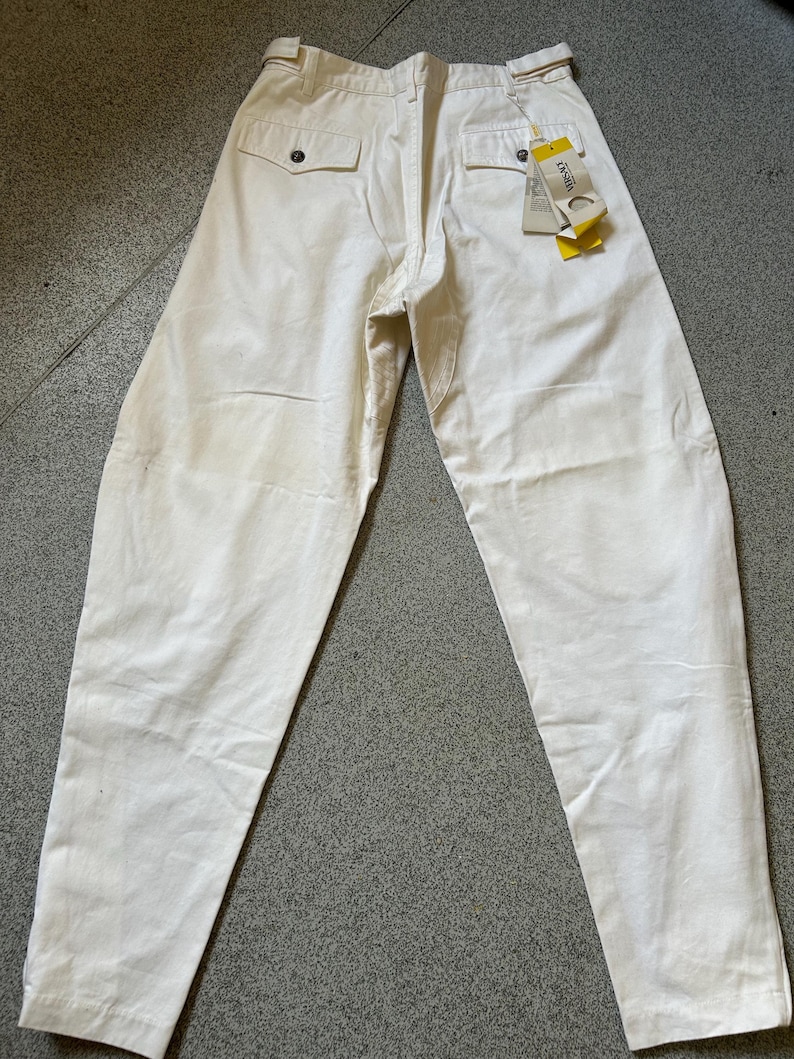 May include: Off-white pants with a relaxed fit, featuring two back pockets with button closures and a belt. A tag with the brand name is attached. The pants are made of a sturdy fabric, suitable for casual wear.