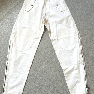 May include: Off-white pants with multiple pockets and knee patches. The pants have black zippers running along the outer seams of the legs. The waist features belt loops and button closures. A tag is attached to the pants.