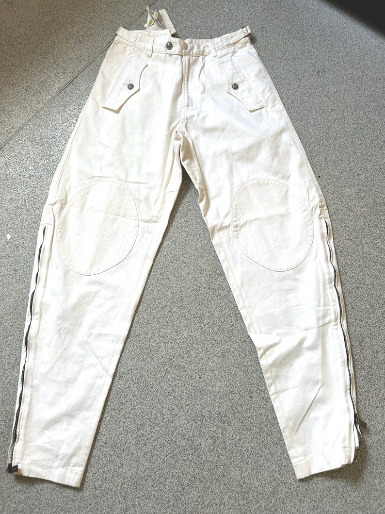 May include: Off-white pants with multiple pockets and knee patches. The pants have black zippers running along the outer seams of the legs. The waist features belt loops and button closures. A tag is attached to the pants.