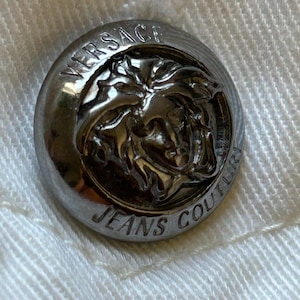 May include: Close-up of a silver Versace Jeans Couture button. The button is round with the brand name and logo embossed on the surface. The button is attached to a white fabric.