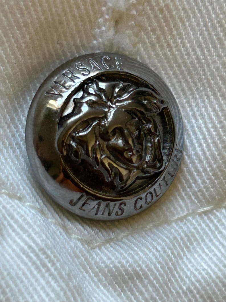 May include: Close-up of a silver Versace Jeans Couture button. The button is round with the brand name and logo embossed on the surface. The button is attached to a white fabric.