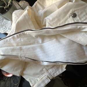 May include: Cream-colored jacket with a black zipper and a button closure. The jacket has a textured, ribbed design on the front and is made of a lightweight fabric. The jacket is folded, showing the interior lining.
