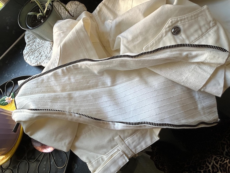 May include: Cream-colored jacket with a black zipper and a button closure. The jacket has a textured, ribbed design on the front and is made of a lightweight fabric. The jacket is folded, showing the interior lining.