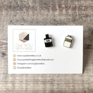 May include: A pair of earrings featuring miniature black and white bottles with the words "GIN" and "TONIC" printed on them.