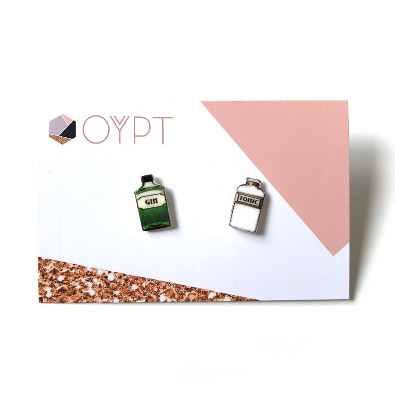 May include: A pair of earrings featuring a miniature green bottle labeled "GIN" and a white bottle labeled "TONIC" on a pink and white card with the text "OYPT".