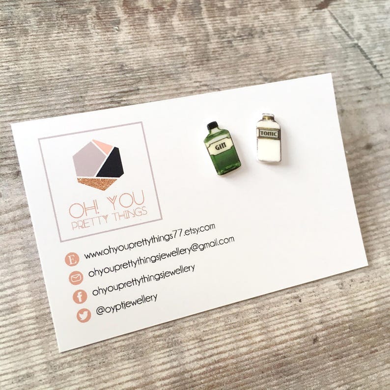 May include: A pair of earrings featuring miniature bottles of gin and tonic. The earrings are made of polymer clay and have a white background with black lettering. The gin bottle is green and the tonic bottle is white.