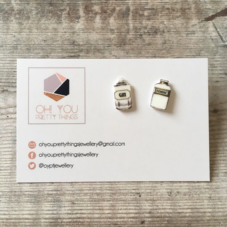 May include: A pair of white and gold earrings shaped like miniature bottles. One bottle says "Gin" and the other says "Tonic".
