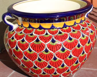 May include: A large, round ceramic planter with a wide opening and two handles. The planter features a vibrant, hand-painted design of red, yellow, blue, and green patterns. The rim is blue, and the interior is white.